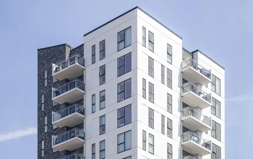 Modern white apartment building with dark brick accents, glass-balconied floors, and large windows under a clear blue sky.