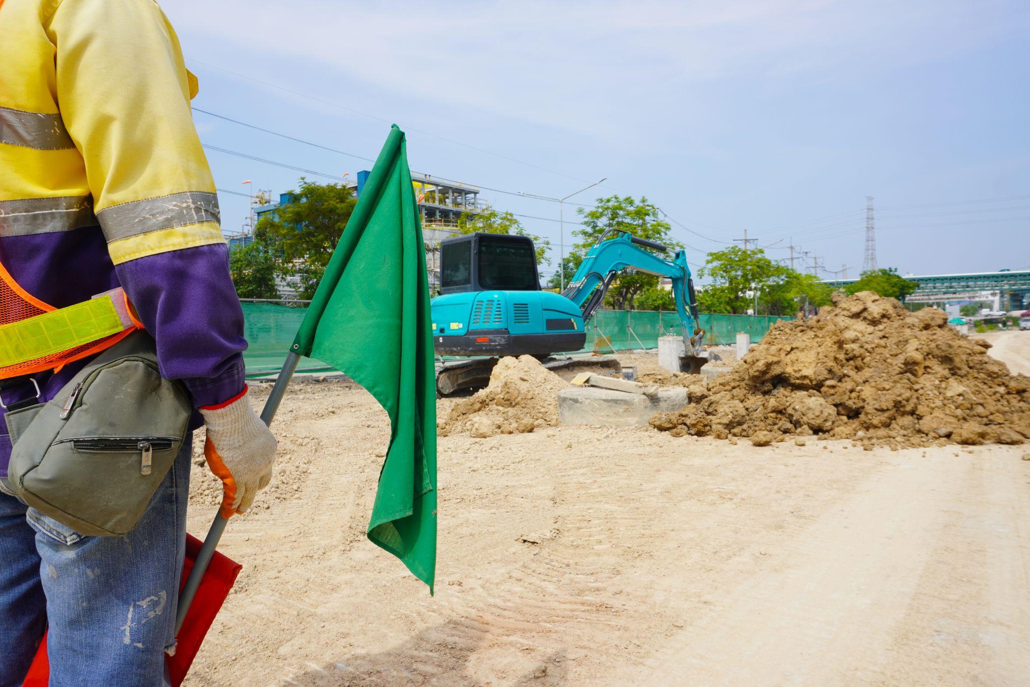 Worker holding a green flag near heavy equipment at an excavation site, representing Maintenance Man Unlimited’s reliable flagging and traffic control services.