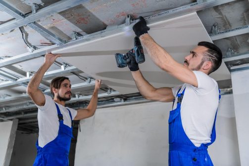 Two construction workers are securing a drywall panel to a ceiling frame